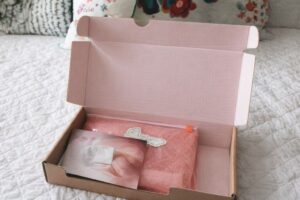 Öppnat paket med rosa modevaror på en vit säng.